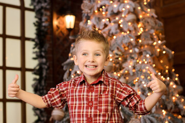 aughing blond boy showing thumbs up with both hands, against the background of New Year's lights garlands on the Christmas tree