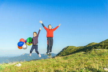 Children jump with balloons in hand that sway in the wind. Mountain peaks and blue sky in background. Children's play, freedom and happiness