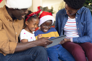 Happy african american family wearing santa hats, using tablet in living room