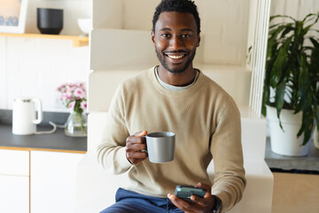 Happy african american man sitting on stairs in kitchen, drinking coffee and using smartphone