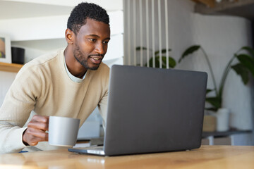 Happy african american man leaning on countertop in kitchen, using laptop