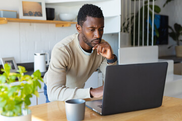 Happy african american man leaning on countertop in kitchen, using laptop