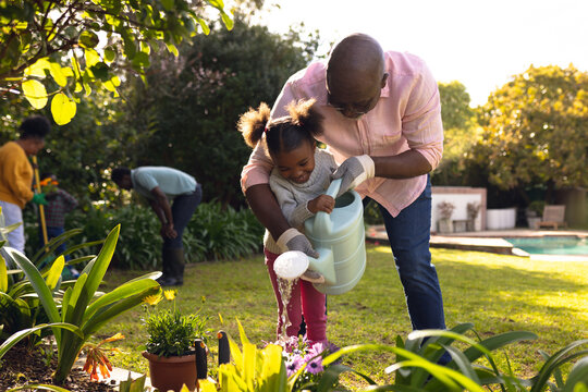 Happy senior african american man and his granddaughter watering plants in sunny garden