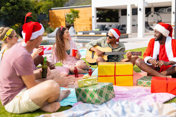 Happy diverse friends with santa hats having picnic in garden at christmas