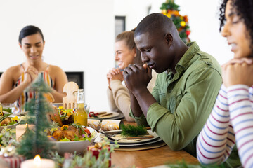 Happy diverse friends sitting at table and praying before dinner at christmas