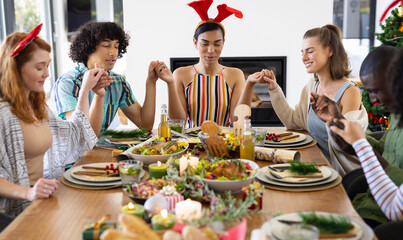 Happy diverse friends sitting at table and praying before dinner at christmas