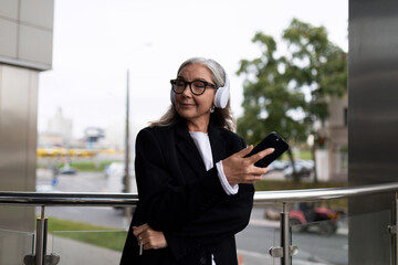 senior stylish woman listening with pleasure to music in headphones by mobile phone in hands outside the office