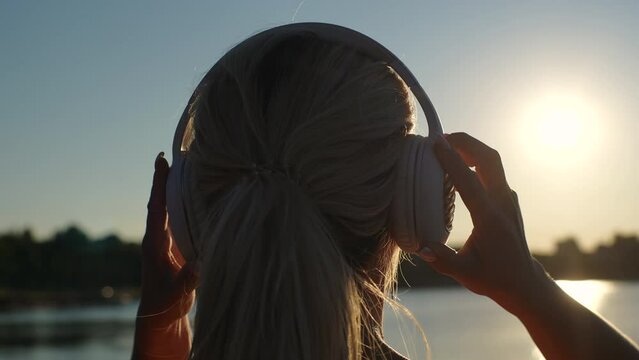 Close-up back view of unrecognizable blonde young woman putting on wireless headphones enjoying music on background of river outdoors at summer morning. Female runner wearing earphones before workout.