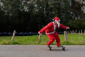 Obraz premium caucasian man dressed as santa claus skating on a longboard skateboard on a country road. christmas and sport concept.