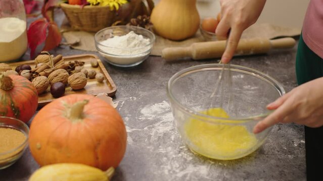 hands cooking pumpkin pie making dough mix eggs and sugar in bowl with whisk. autumn seasonal decoration fall orange pumpkins brown walnuts on kitchen table flour dough yellow pastry thanksgiving day