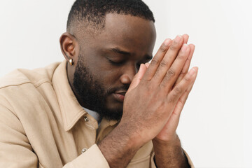 Fototapeta premium African American believing man sitting at home on sofa praying to God, asking for good luck, thoughtful bi-racial superstitious man holding hands in prayer, feeling gratitude, faith, religion concept