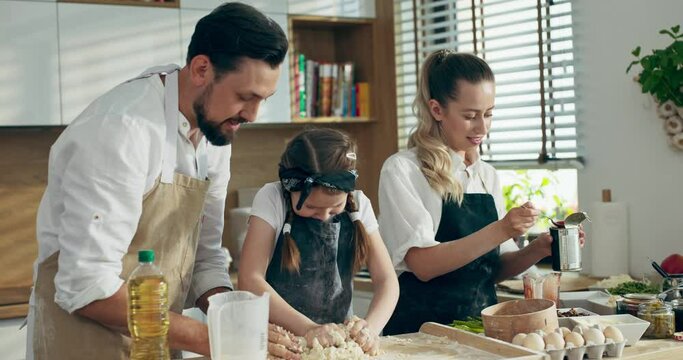 Happy family concept. Delighted mother and father teaching preschooler kid girl kneading dough. Adorable wife preparing tomato sauce while father with child preparing homemade dough for pizza.