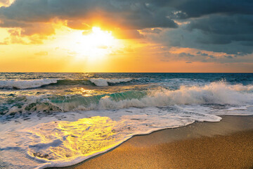 Beauty of sea nature of Mediterranean coast on warm summer evening. Setting sun illuminates stormy waves with caps of foam rolling onto golden sandy beach.