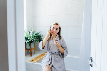 Young woman in bathrobe looking in the mirror and applying facial natural cosmetic clay mask on her face in bathroom. Cosmetic procedures for skin care at home. Beauty self-care. Selective focus.