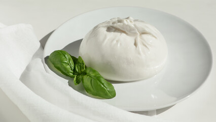 Traditional Italian, fresh, soft burrato ball of young mozzarella cheese with cream on a round white plate with basil greens on a light background. close-up