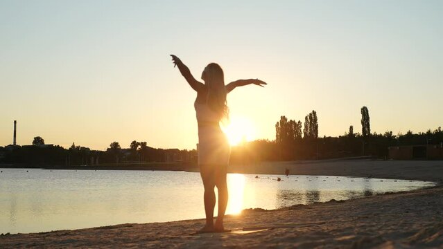 Silhouette of fit young woman standing on Sadhu nail board raising arms to sky on sandy beach on background of sunlight on summer morning. Inspired female practicing yoga exercise at seascape.