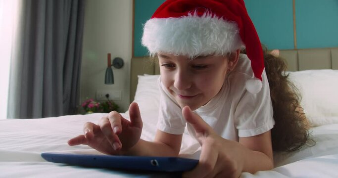 Child with digital tablet. Kid dream online video games at Christmas. Daughter kid in Christmas hat Santa Clausa watching online video under covers with digital tablet network. child on social media