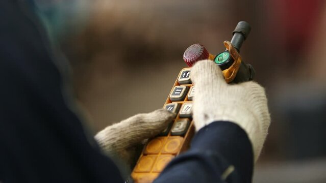 A pair of gloved hands of a manual worker pressing the direction button on an industrial yellow remote. It controls a load of metal bars tied up in a bundle and being hoisted in the direction 