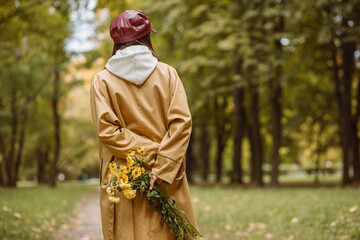 Back view of woman in trench with bouquet of yellow flowers in hands behind back walking along path...