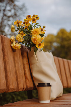 Blank paper cup of takeaway coffee with flower bouquet in bag placed on bench in autumn park 
