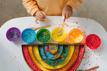 Child hands playing colored rice and make rainbow. Child filled the rainbow with bright rice. Montessori material. Sensory play and learning colors activity for kids. - obrazy, fototapety, plakaty