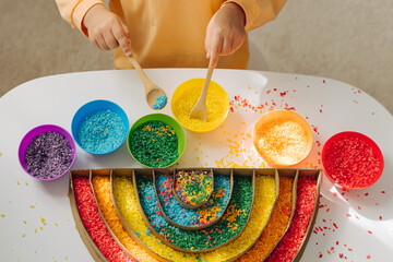 Child hands playing colored rice and make rainbow. Child filled the rainbow with bright rice. Montessori material. Sensory play and learning colors activity for kids.