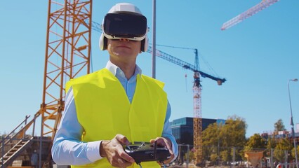 Professional drone operator in virtual reality helmet standing in front of construction site. Builder holding remote controller. Office building and crane background. © Acronym