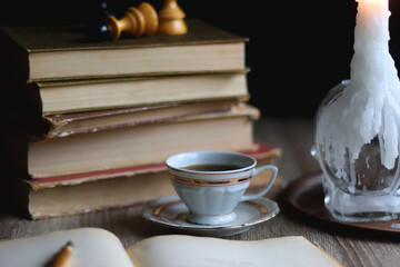 Stack of vintage books, cup of tea or coffee, lit candles, reading glasses and chess pieces on wooden table. Dark academia concept. Selective focus.