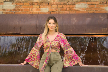 Young, attractive and blonde woman, wearing cashmere shirt and green trousers, sitting on a water...