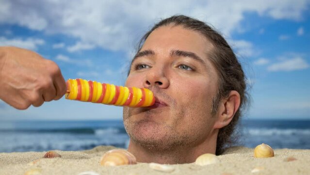 A man buried in sand on beach licking a lolly