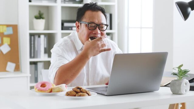 Asian fat man eating donut, sweet, junk food during working with computer laptop, unhealthy eating concept