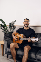 A young white hipster male with glasses, beard and tattoos in black t-shirt sitting on a grey sofa...