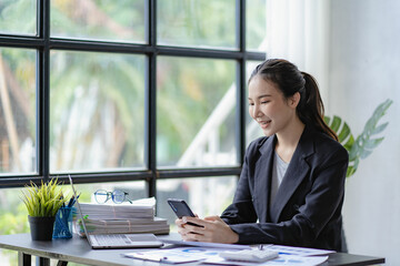 Asian businesswoman using smartphone to talk to customers and calculate budget Company expenses and income from charts, reports, documents, budget plans, finance, accounting, laptop analysis.