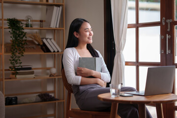 Happy Asian female college student reading book and working on her laptop in the living room during the holidays. Asian woman reading book at home.