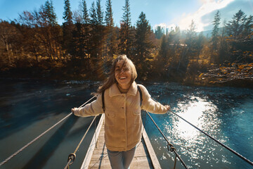 Wide angle view of happy smiling backpacker woman looking to camera at sunny day, standing on suspended bridge across river