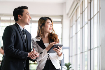 Senior Asian businessman and young female businesswoman discuss in the office, working on social media strategy, looking outside with blur colleagues in the background. Image with copy space.