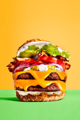 Enorme burger au boeuf, bacon et cheddar avec tomates et pickles cornichons, oignons, laitue photographié en studio sur fond jaune et vert.