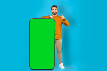 A man in a yellow shirt standing near a huge smartphone with a blank screen and pointing his thumb at it, mockup, copy space, advertising concept