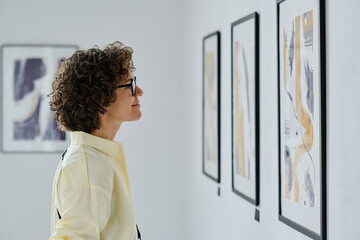 Young woman in eyeglasses standing in front of pictures on the wall, examining the art and smiling