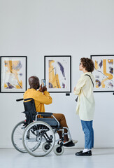 Rear view of African man in wheelchair making photo on mobile phone while visiting art gallery together with woman