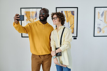 Young multiethnic couple taking selfie on smartphone while visiting art gallery