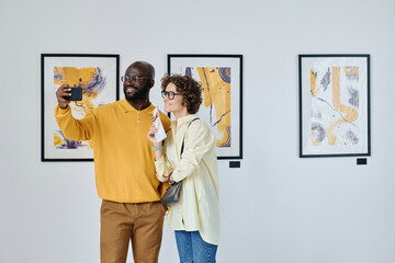 Multiethnic couple of visitors taking selfie on mobile phone at art gallery