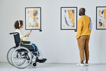 Young woman in wheelchair listening to guide with man standing near by while they visiting art gallery
