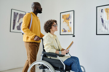 Woman with disability enjoying modern art at gallery with African man helping her to push wheelchair