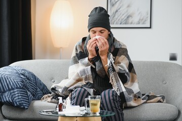 Sick sad man sits on couch at home suffers from runny nose flu disease coronavirus pandemic covid...