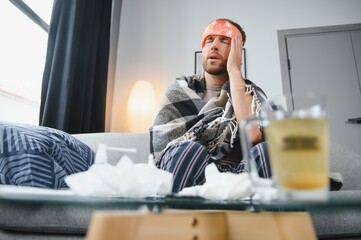 Sick bearded man who has bad cold or seasonal flu sitting on couch at home. Guy with fever wearing...