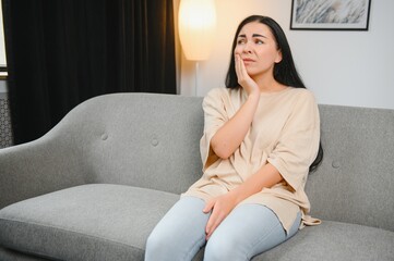 Woman with severe toothache touching her swollen cheek. Young girl feeling discomfort after tooth extraction. Upset lady with inflamed tooth nerve or terrible dental pain sitting on sofa at home.