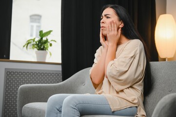 Obraz premium Woman with severe toothache touching her swollen cheek. Young girl feeling discomfort after tooth extraction. Upset lady with inflamed tooth nerve or terrible dental pain sitting on sofa at home.