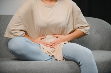 Fototapeta premium Young female suffering form stomach ache while sitting on couch at home. Causes of abdominal pain include menstruation pain, gastritis, stomach ulcer, food poisoning, diarrhea or IBS. Selective focus