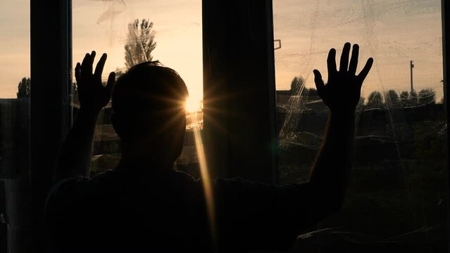 A sad man looks out the window at sunset during the war in Ukraine. A young man in front of sealed windows from a blast wave of rockets. War man in depression at home.